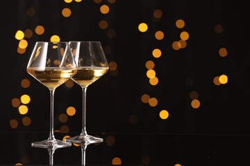 Tasty white wine in glasses on mirror surface against dark background with blurred lights, space for text. Bokeh effect