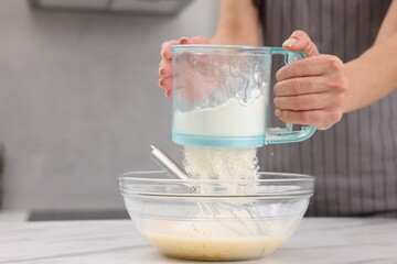 Woman adding flour into bowl with dough at white marble table, closeup