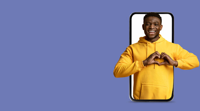 A young man wearing a yellow hoodie smiles as he creates a heart shape with his hands while interacting with a large smartphone screen. The background is a solid color, enhancing the focus on him.