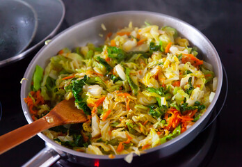 Chopped cabbage braising with vegetables in open pan. Wooden spatula for stirring.