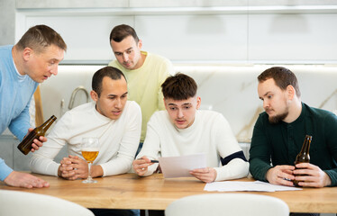 Upset worried man sitting at table in home kitchen with papers in hands, reading unexpected medical results with group of male friends solacing and comforting him ....