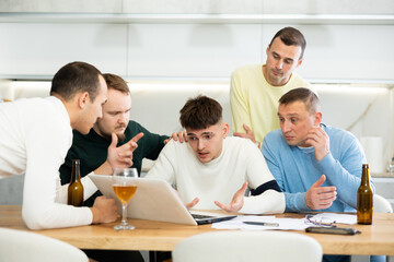 Obraz premium Group of men huddling around laptop at kitchen table, discussing problems and comforting distressed friend while gathering at home bachelor party