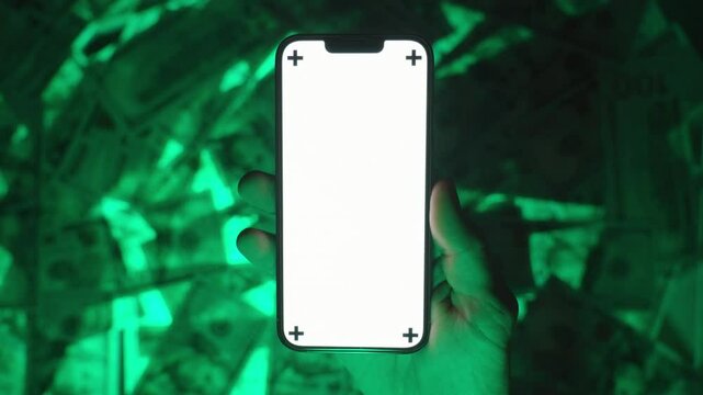 Close-Up of a Hand Holding a Smartphone with a Blank White Screen Mockup and Tracking Markers, Displayed Against a Green-Glowing Background of US Hundred-Dollar Bills, Highlighting Technology and Fina