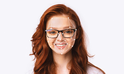 A young woman with red hair and glasses smiles while facial recognition technology tracks her...