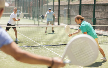 Adult man and elderly man doubles playing against two men in padel tennis on court