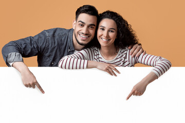 Happy Young Arab Couple Pointing Down At Big White Advertisement Board, Smiling Middle Eastern...