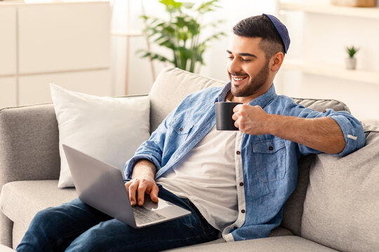 People And Technology Concept. Portrait of smiling young jewish man in kipa using laptop holding cup and drinking hot coffee. Male sitting on couch watching video, movie or webinar, taking break