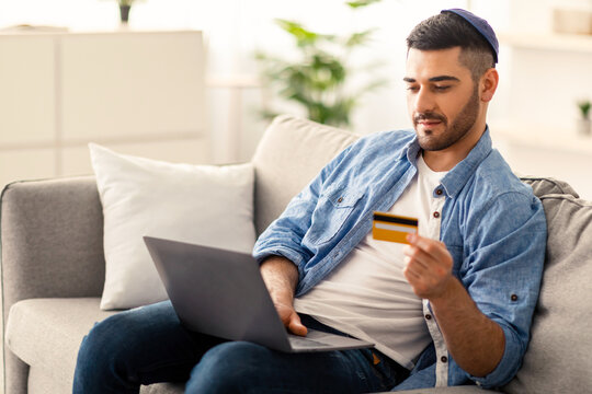 Electronic Payment Service Concept. Portrait of casual young jewish businessman in kippa holding credit card and using laptop, making e-payments while working with computer at home office, copy space