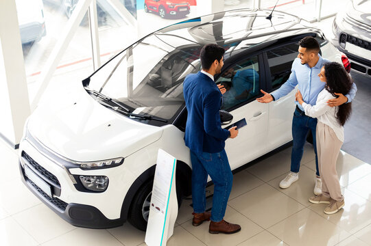 Top view of middle eastern couple choosing new auto at car showroom, young man and woman embracing while having conversation with male sales manager, standing by nice white automobile - Powered by Adobe