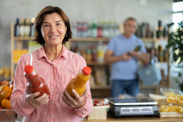 Fototapeta premium Mature woman hold plastic bottles, choose consider juice in store, man customer in background. Clients look package of fruit juice, inspect product, reflect and consulting before buying.