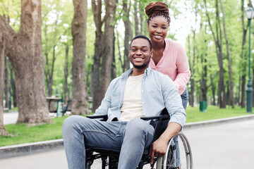 Fototapeta premium Cheerful young black couple with lovely woman and millennial guy in wheelchair going for walk at park, spending spring morning outdoors, full length portrait. Copy space