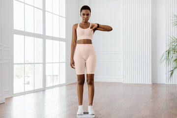 Fototapeta premium Full Body Length Of Sad And Disappointed African American Woman Standing On Bathroom Scales, Unhappy About Her Body Parameters, Showing Thumb Down Sign Gesture. Slimming Fail, Unsuccessful Weight Loss