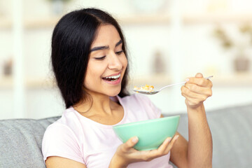 Balanced nutrition concept. Lovely Indian lady eating delicious cereal with milk on cozy couch at home. Beautiful Eastern woman leading healthy lifestyle, having wholesome meal indoors