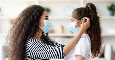 Side view of young brunette woman putting face mask on her cute curly daughter, home interior....
