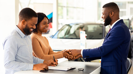 Buying Auto. Grateful Black Spouses Handshaking With Manager In Dealership Center After Purchasing New Car, African American Couple Signing Papers In Modern Automobile Showroom