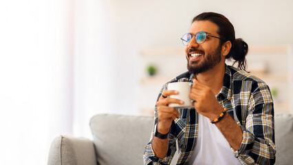 Cheerful young indian man with coffee mug looking at copy space, sitting on couch in cozy living...