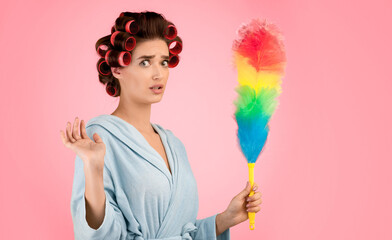 Spring Cleaning. Confused Glamorous Lady With Hair Curlers Holding Feather Duster Doing House Chores Looking At Camera In Shock Standing Over Yellow Studio Background. Housework Concept