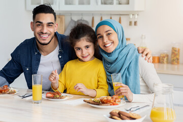 Portrait Of Happy Muslim Family Of Three Posing At Camera While Eating Breakfast In Kitchen Together, Cheerful Middle Eastern Parents And Their Little Daughter Sitting At Table Enjoying Tasty Food