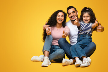 We Recommend. Portrait Of Happy Arab Family Of Three Showing Thumbs Up At Camera, Joyful Middle Eastern Parents And Little Daughter Gesturing Sign Of Approval, Posing On Yellow Background, Copy Space