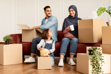 Real Estate Purchase, House Move, Relocation Concept. Portrait Of Cheerful Muslim Family Unpacking Cardboard Boxes, Sitting With Open Packages On The Couch And Floor In Living Room. Housing
