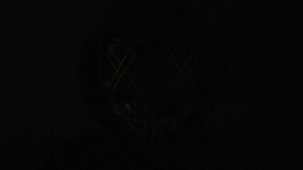 A hooded figure with a glowing green neon mask moves through the darkness, red and blue strobe lights flashing around him. The flickering glow heightens the tension, creating a chilling atmosphere for - Powered by Adobe