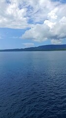 view in the middle of the blue sea during the day with a background of hills and white cloudy skies