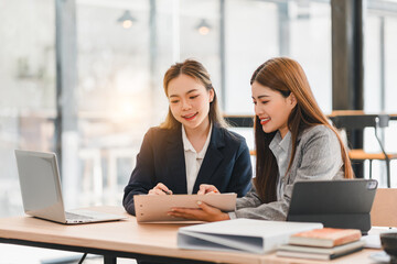 Fototapeta premium Two businesswomen collaborating in modern office, discussing work on tablet while surrounded by laptops and documents, showcasing teamwork and productivity