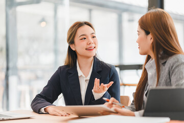 Two professional women in business attire having friendly discussion at modern office table, with laptop and documents, showcasing collaboration and teamwork