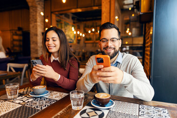 Portrait of happy couple sitting next to each other with mobile phones in hands and texting messages