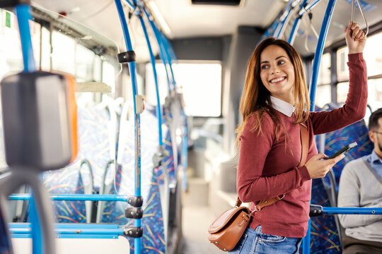Portrait of happy female commuter commuting by city bus while standing and using phone.
