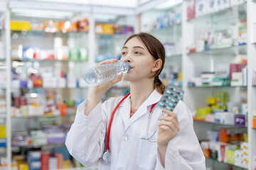 woman pharmacists drinking water holding capsule blister pack at shelf stocked in pharmacy drugstore.