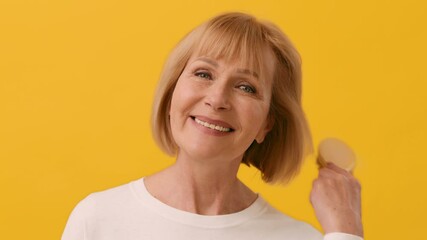 A woman with a bob haircut gently brushes her hair with a wooden hairbrush in a cheerful yellow setting, showcasing a moment of self-care and grooming joy.