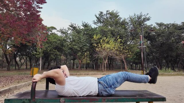Young Man Performing Lying Leg Raises for Lower Abs on a Bench in the Park, with Trees, Evening Light, and Cars in the Background, in Slow Motion