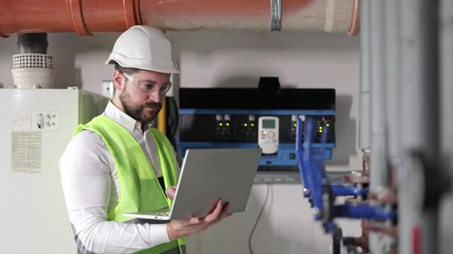 Serious bearded man engineer in glasses, safety helmet, and vest holds laptop types, working in boiler room. Focused male mechanic checks heating system equipment.