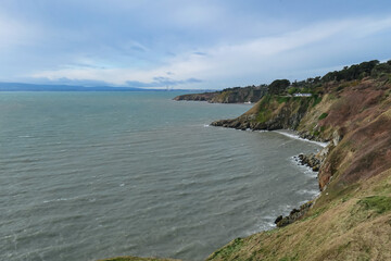 Fototapeta premium Rugged coastline in Howth, Dublin, Ireland under grey overcast sky. Steep cliffs covered in dry vegetation drop down to choppy Atlantic Ocean. Wild windswept beauty. Peaceful solitude, coastal beauty