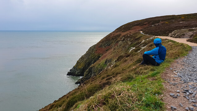 Man on scenic path winds along steep, grassy cliff overlooking grey sea under overcast sky. Coastline in peninsular village Howth in Dublin, Ireland. Peaceful solitude and coastal beauty, Irish coast