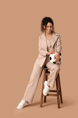 Female African-American soccer trainer in business suit with ball sitting on chair against beige...