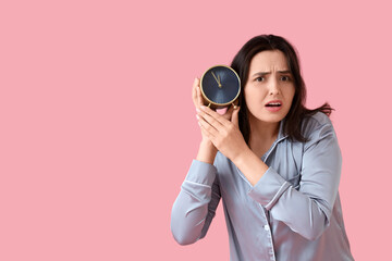 Stressed young woman in pajamas with alarm clock on pink background