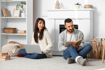 Loving young happy couple with laptop and book sitting near fireplace at home