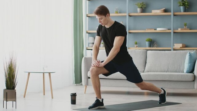 A young Caucasian man engages in a lunging exercise at home, showcasing his athletic physique. The room features modern decor with a plant and a stylish couch.