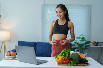 Asian nutritionist is measuring her waistline with a measuring tape during a video call on her laptop, showing the results of her healthy diet with fresh vegetables and fruits on the table