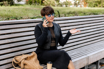 Brunette with short hair, sitting on a bench in a city park, holding a phone, talking,...