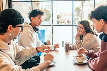 Four diverse young students are sitting at a table in a cafe, enjoying coffee and engaging in...