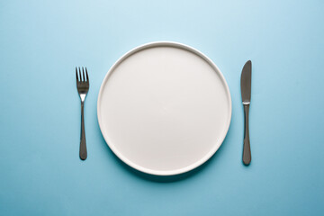 Top view of empty ceramic white plate and cutlery on blue background
