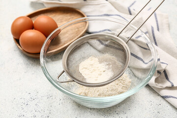 Metal sieve with flour in bowl, chicken eggs and napkin on light background