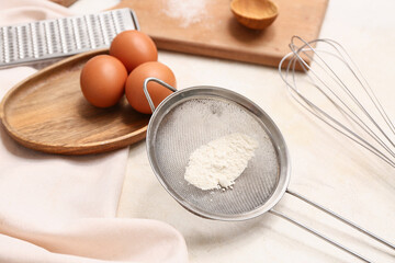 Metal sieve with flour, whisk, chicken eggs and grater on light background