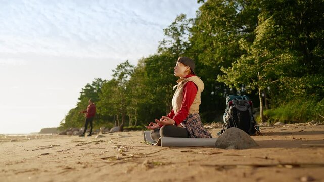 Enjoy silence and calmness, woman tourist meditating on sea shore after hiking. Halt on coastline after traveling across forest, woman backpacker practicing yoga and breathing exercise fore recovery