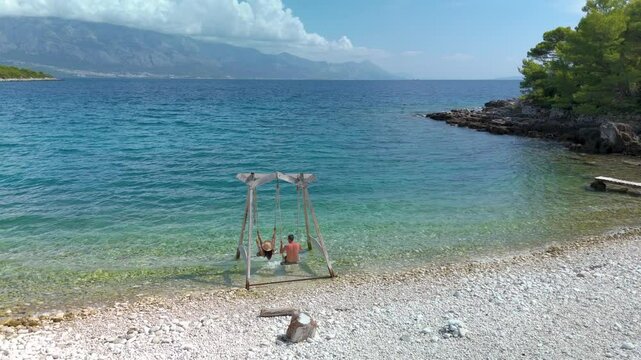 Enjoy the tranquil moment as two friends sway gently on a seaside swing, surrounded by crystal-clear waters and stunning Croatian landscapes under a bright blue sky at brac Island