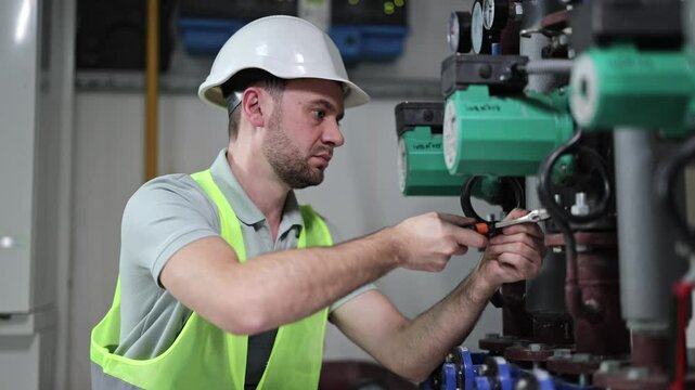 Focused male engineer in boiler room examining heating system equipment. Professional worker in hardhat fixing pipes in power generation plant.