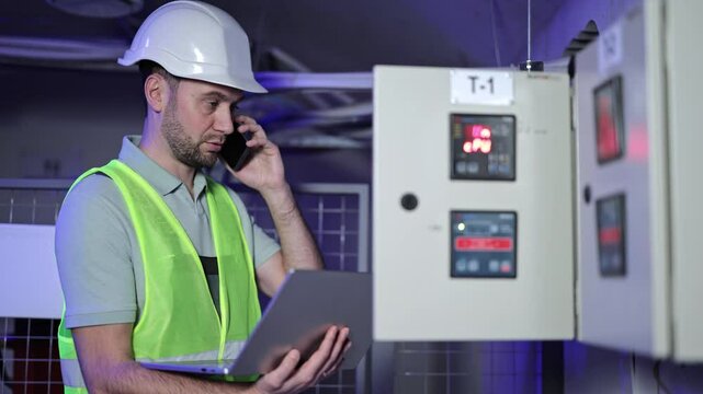 Serious electrical engineer talking on phone and holding laptop, inspects electrical control panel in server room. Focused maintenance technician monitoring equipment.